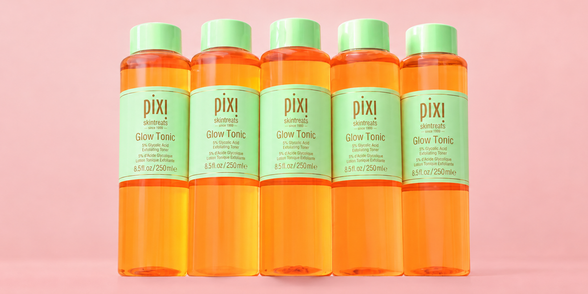 Five Pixi Glow Tonic bottles filled with bright orange liquid, arranged in a row against a solid pink background. View 1