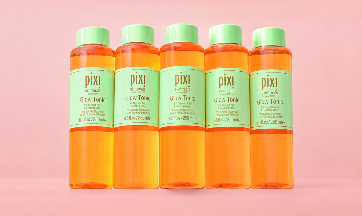 Five Pixi Glow Tonic bottles filled with bright orange liquid, arranged in a row against a solid pink background. View 2