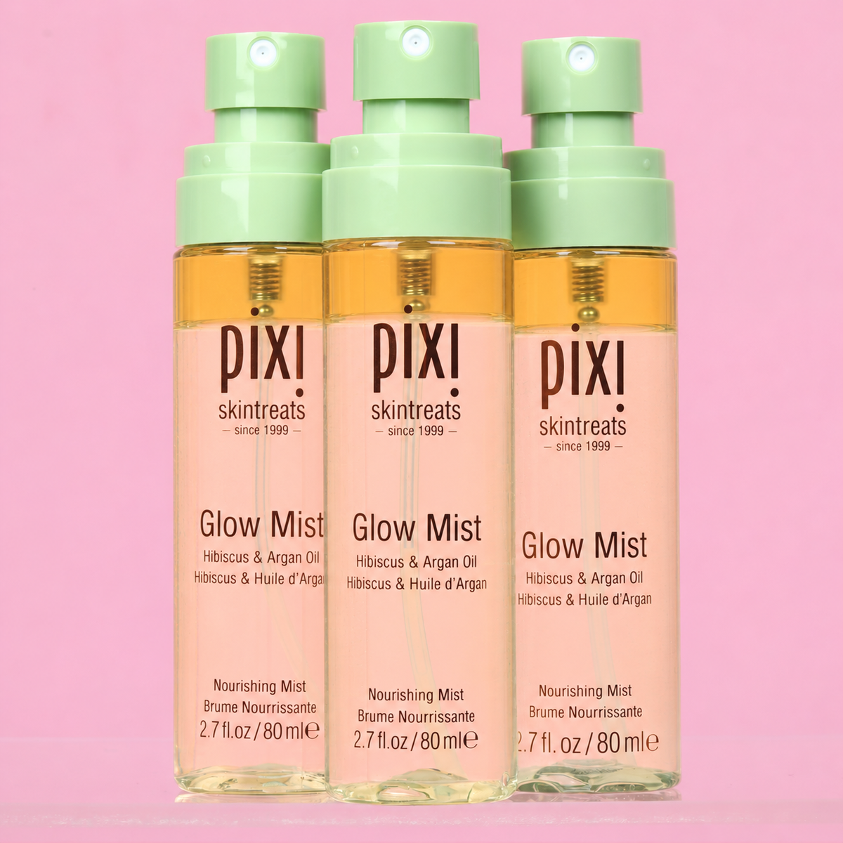 Three Pixi Skintreats Glow Mist bottles with pale green pump tops, filled with a warm golden liquid, set against a solid pink background.