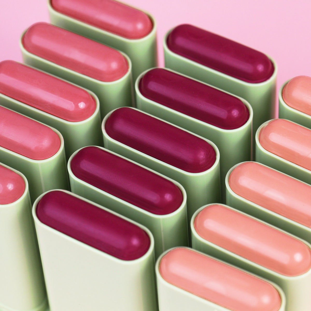 A close-up arrangement of Pixi On-the-Glow Blush sticks Cassis, Mauve and Chantilly set against a solid pink background.