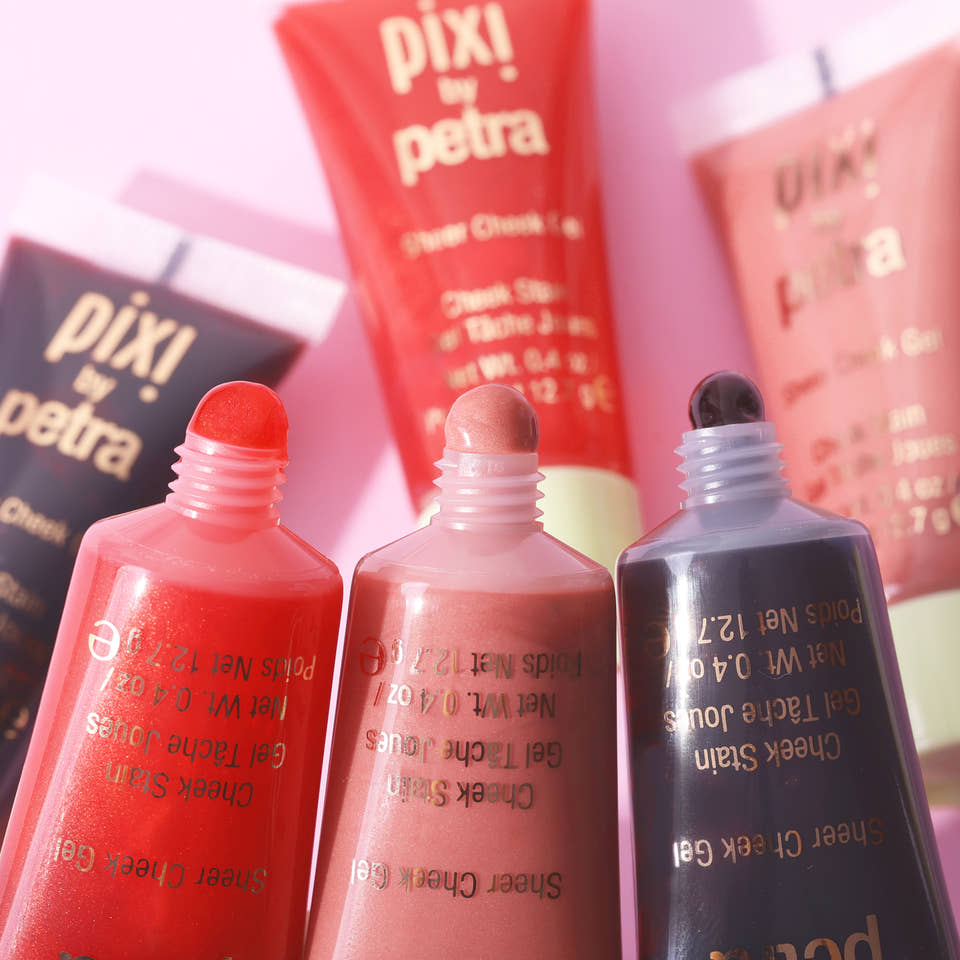 Three open Sheer Cheek Gel tubes of Pixi by Petra cheek stain in shades of coral red, nude pink, and dark charcoal, arranged in a close-up studio shot against a soft pink background. view 2