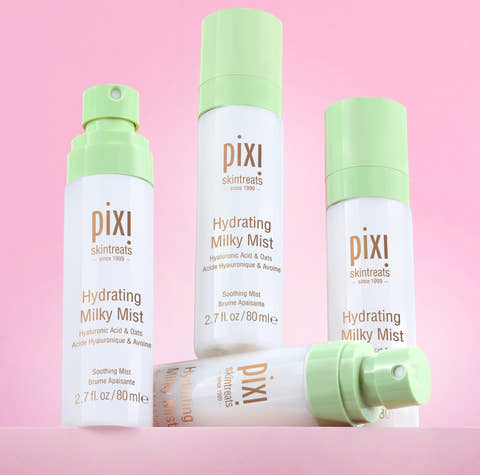 Three Pixi Skintreats Hydrating Milky Mist bottles in white and light green packaging, arranged on a solid pink background with one bottle lying down in front.